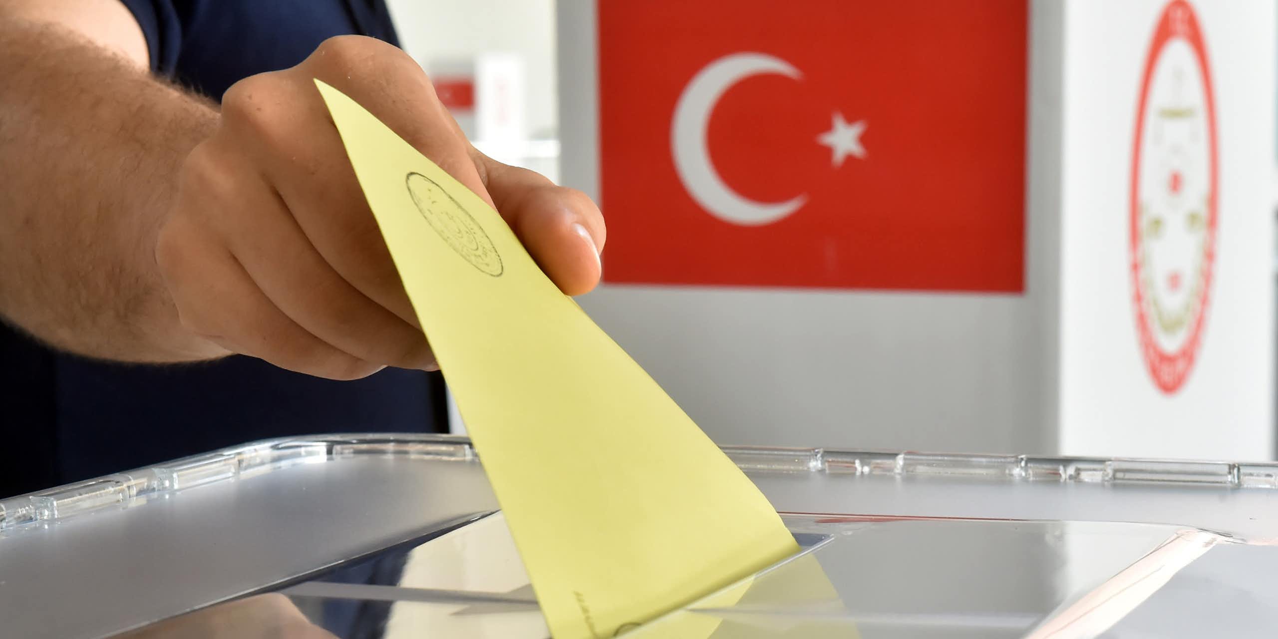A man puts a ballot into a ballot box in front of the national flag of Turkey.